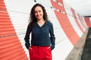 A woman engineer stands in front of sheet metal with her hands in her pockets and smiling
