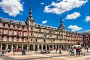 A bustling plaza with people walking and cycling past, capturing the vibrant spirit of a 2023 event in front of an ornate, multi-story building. The blue sky with scattered clouds adds charm to this lively scene.