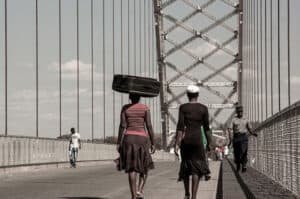 Women walking across a bridge, one of them has a luggage bag balancing on her head