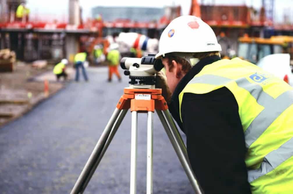A man in a hard hat and high visibility vest looks through a surveying camera device on a tripod