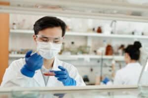Man wearing a lab coat, mask, gloves and safety goggles drops a sample into a petri dish