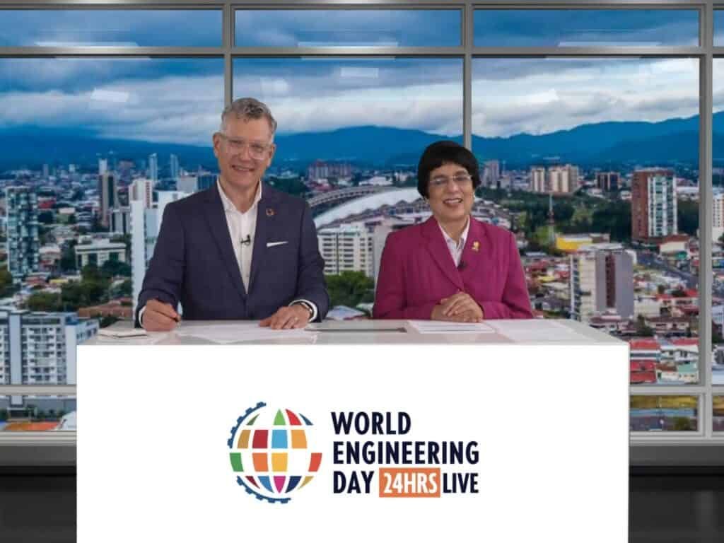 Two people sit at a desk with the "World Engineering Day 24HRS LIVE" sign, embodying the spirit of celebration. A stunning cityscape unfolds through the large windows behind them.