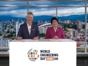 Two people sit at a desk with the "World Engineering Day 24HRS LIVE" sign, embodying the spirit of celebration. A stunning cityscape unfolds through the large windows behind them.