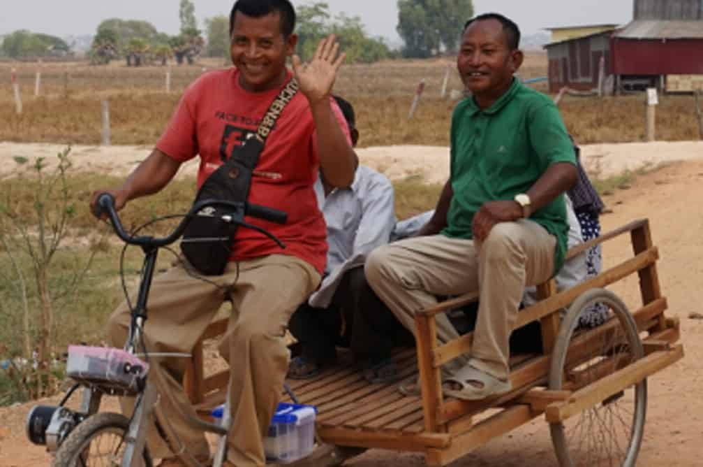 Two men on a tuk tuk, one is waving at the camera