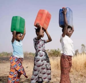 Three individuals carrying large containers on their heads navigate a dry, grassy area, reminiscent of the teamwork and endurance often seen at a hackathon.