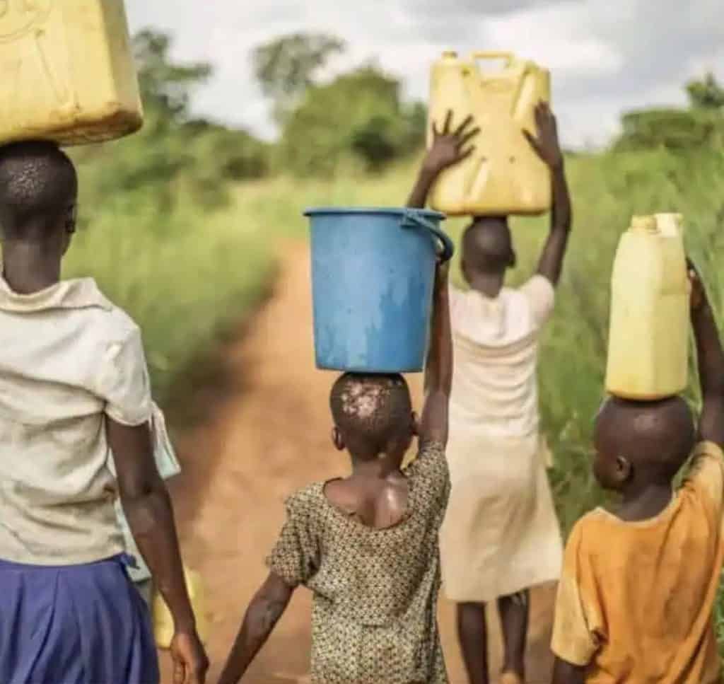 Four people walk down a dirt path, each balancing a container on their head, in a rural setting.