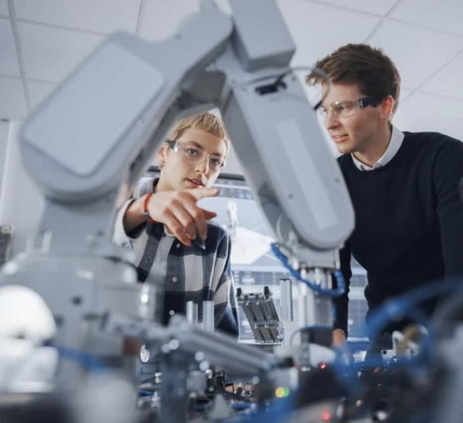 Two people in safety glasses examine equipment in a laboratory setting, focusing intently on a robotic apparatus.