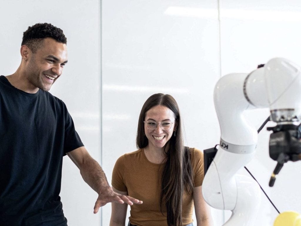 Two people stand beside a robotic arm, both smiling. The man gestures toward the robot while the woman observes, celebrating their innovation on World Engineering Day in a bright, indoor setting.