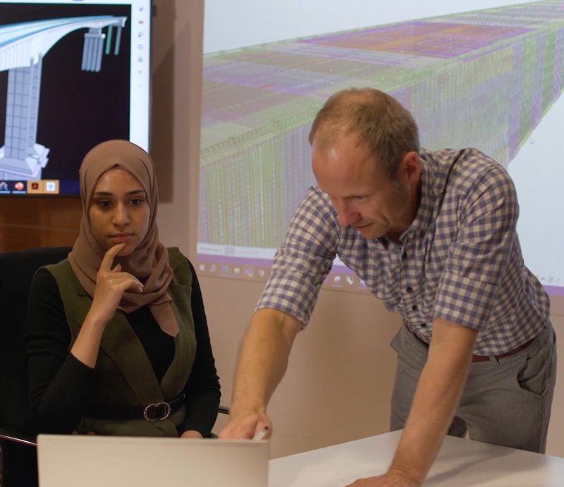Two people in an office setting. A woman sits and observes as a man leans over to point at a laptop. A large screen displays a technical diagram in the background.