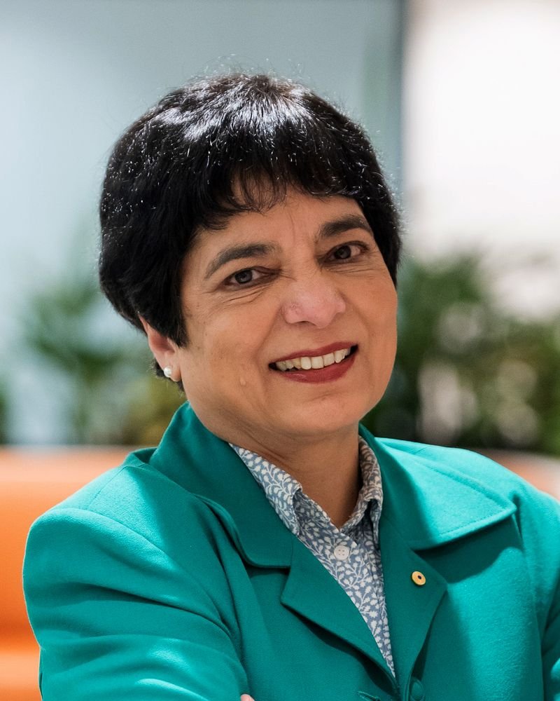 A person with short black hair, wearing a green blazer and a patterned shirt, reminiscent of Marlene Kanga's style, is smiling with arms crossed in an indoor setting.