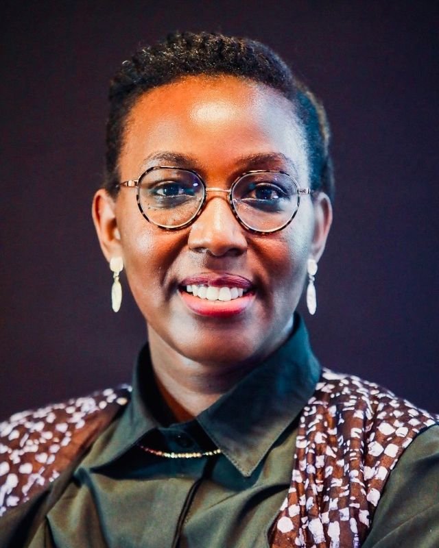Lydie E. Uwantege, wearing glasses and earrings, smiles warmly in a black shirt and patterned vest against a dark background.