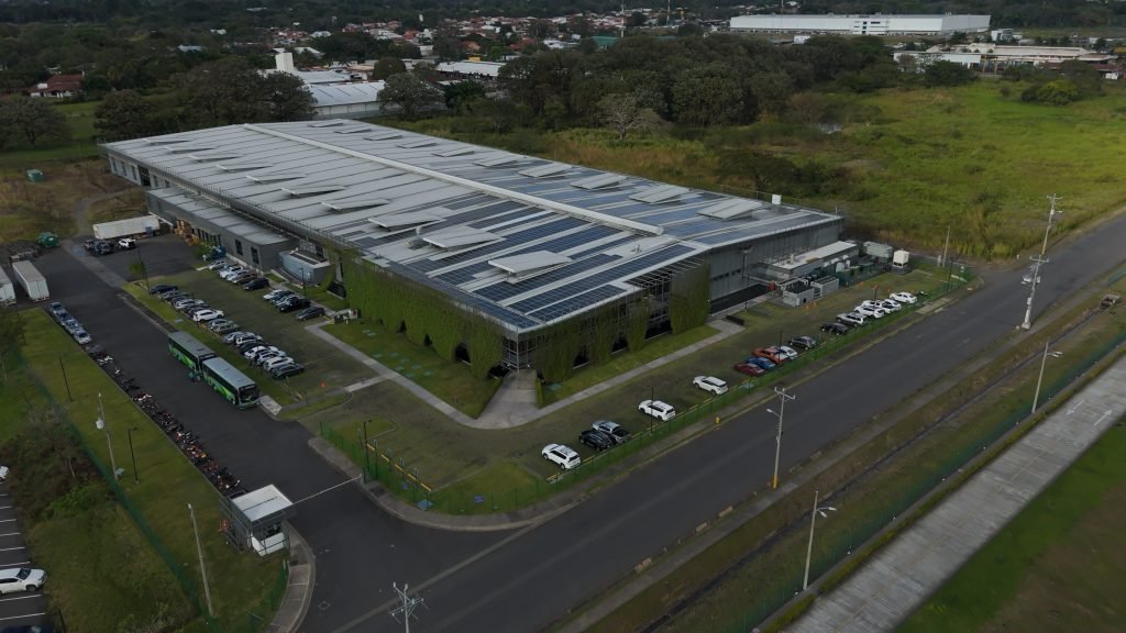 Aerial view of a large warehouse-style building with solar panels on the roof, surrounded by parking lots, greenery, and roads.
