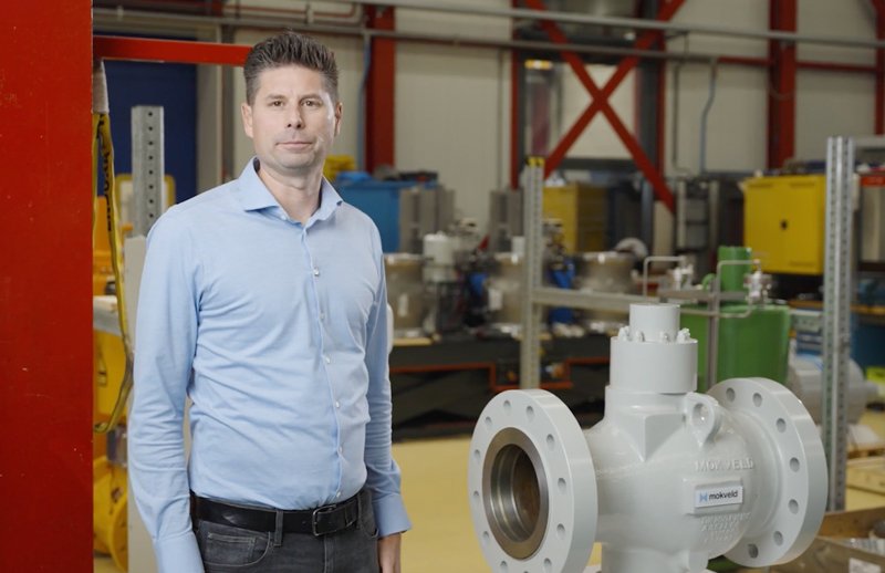A man in a light blue shirt stands next to a large industrial valve in a workshop setting.