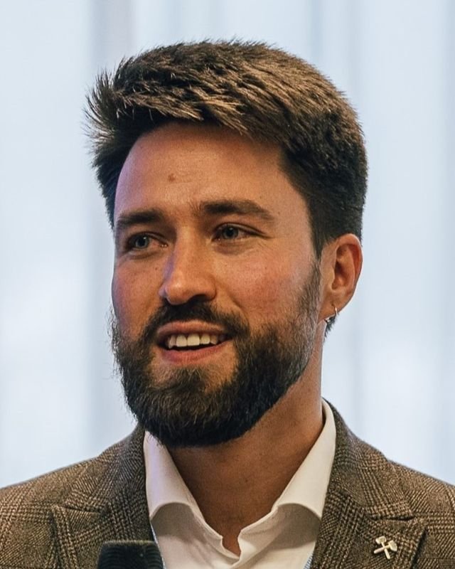 Dr. Alexey Alekseenko, a distinguished man with a beard and earring, is engaging in conversation while dressed in a white shirt and brown jacket.