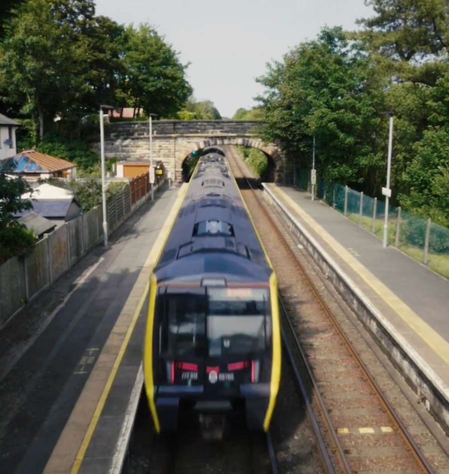 A train swiftly maneuvers through a UK station with two platforms, set in a green, tree-lined area under a stone bridge on a clear day, exemplifying efficient transport.