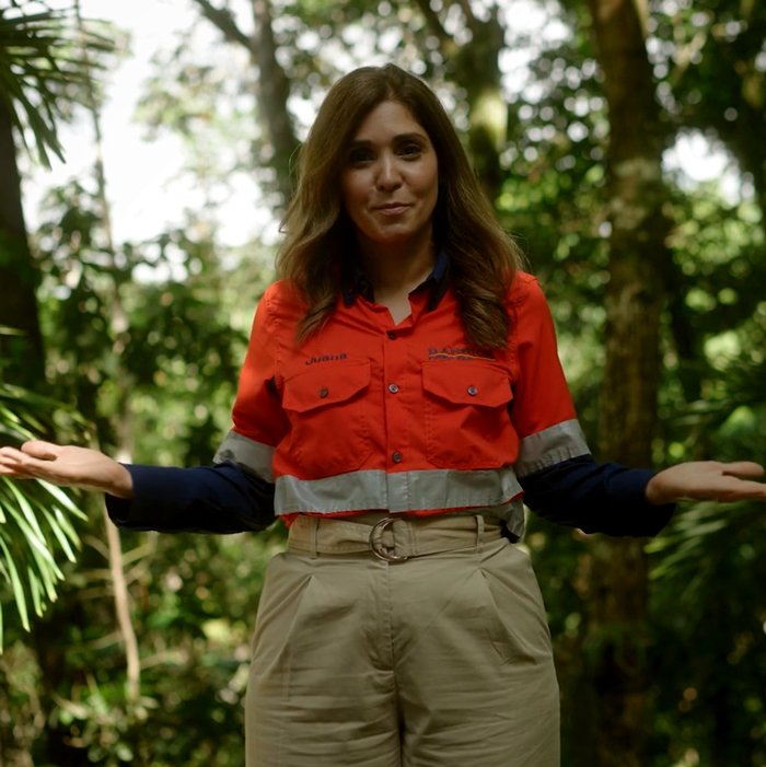 A person in an orange work shirt and khaki pants is standing in a forested area with arms slightly outstretched.