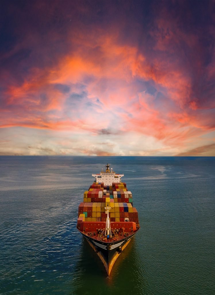 A large container ship sails towards the camera under a vibrant, cloudy sunset sky over calm waters.