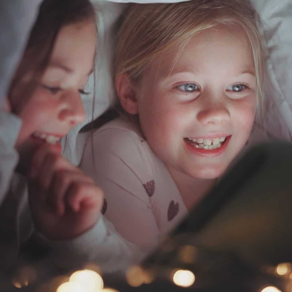 Two children under a blanket, smiling and looking at a lit device. Fairy lights are visible in the foreground.