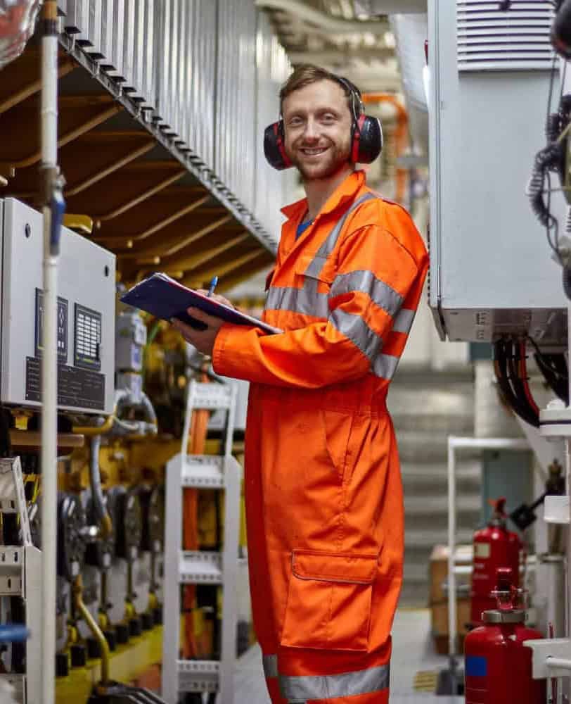 Person in an orange safety suit and ear protection holding a clipboard, standing in an industrial setting with machinery in the background.
