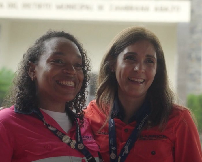 Two women smiling, wearing collared shirts with logos and lanyards, standing side by side in an outdoor setting.