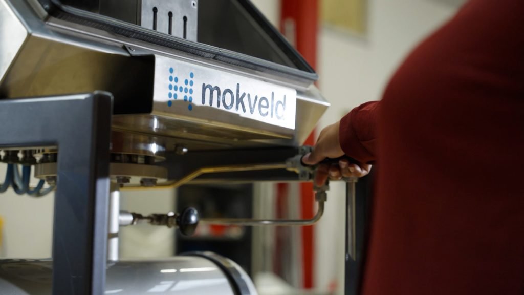 A person operates a control mechanism on a machine with a "mokveld" logo visible on the casing.