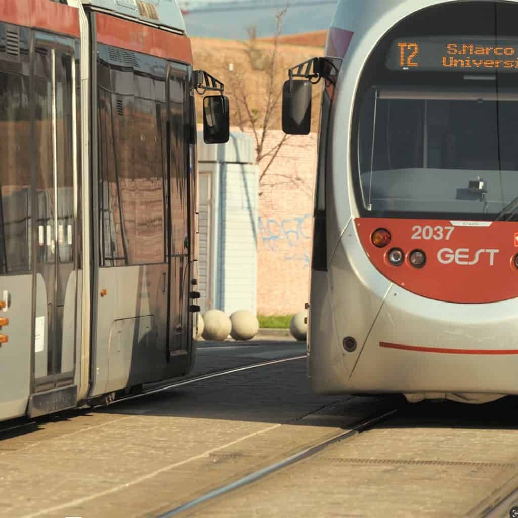 Two trams facing each other on parallel tracks. The tram on the right displays route "T2 S. Marco - Universita" and number "2037." The setting is urban with a brick wall nearby.