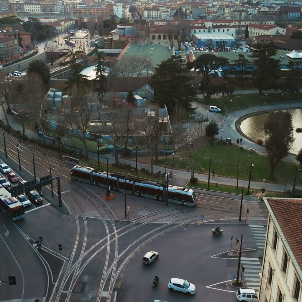 Aerial view of a city intersection with a tram, cars, and a park with a pond. Buildings and trees are visible in the background.