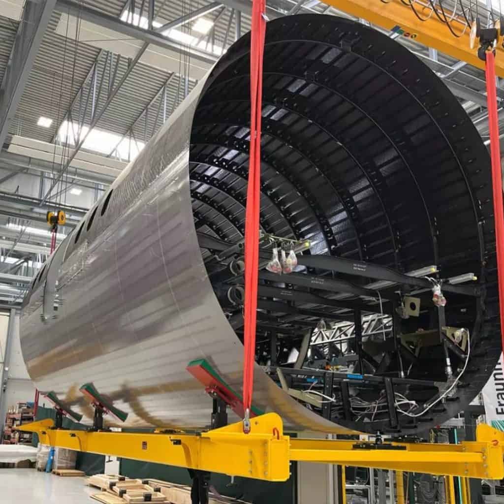 Large cylindrical metal structure suspended in a fabrication facility, supported by red straps attached to a yellow crane.