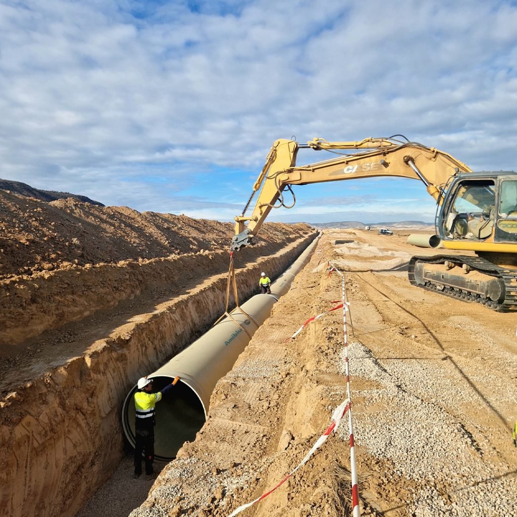 Construction site with an excavator lowering a large pipe into a trench. Workers in safety gear are guiding the pipe. Desert landscape and cloudy sky in the background.