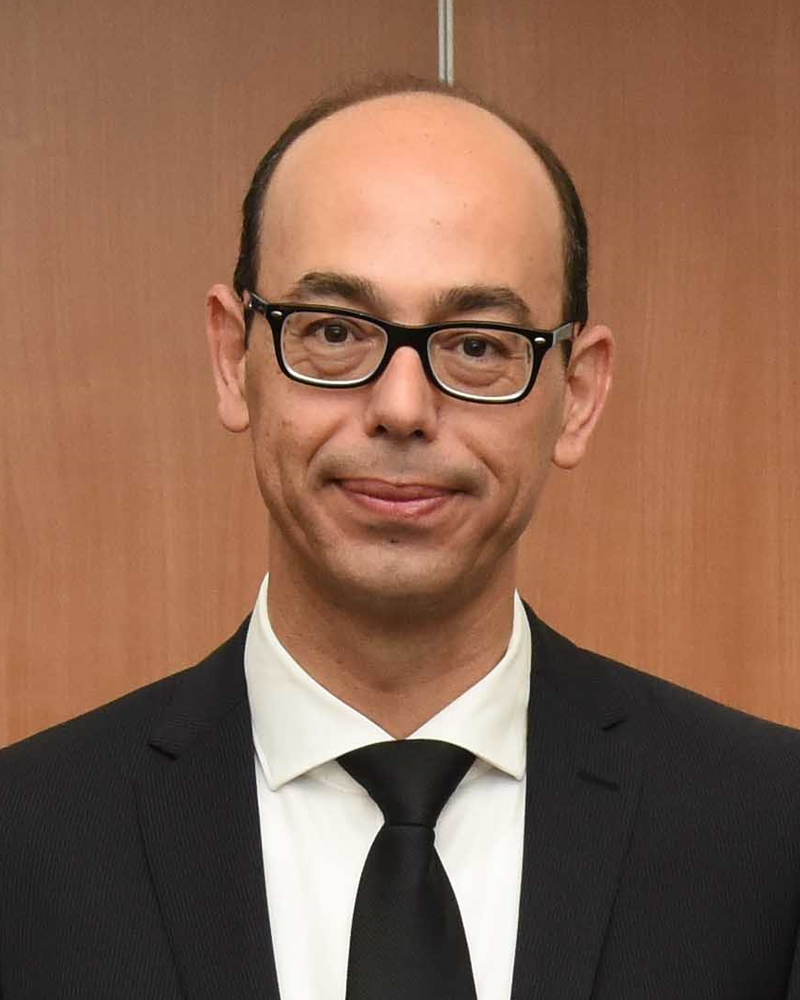 A man in a black suit and tie, reminiscent of Platonas Stylianou's classic style, stands before a wooden background, his glasses reflecting a hint of intrigue.