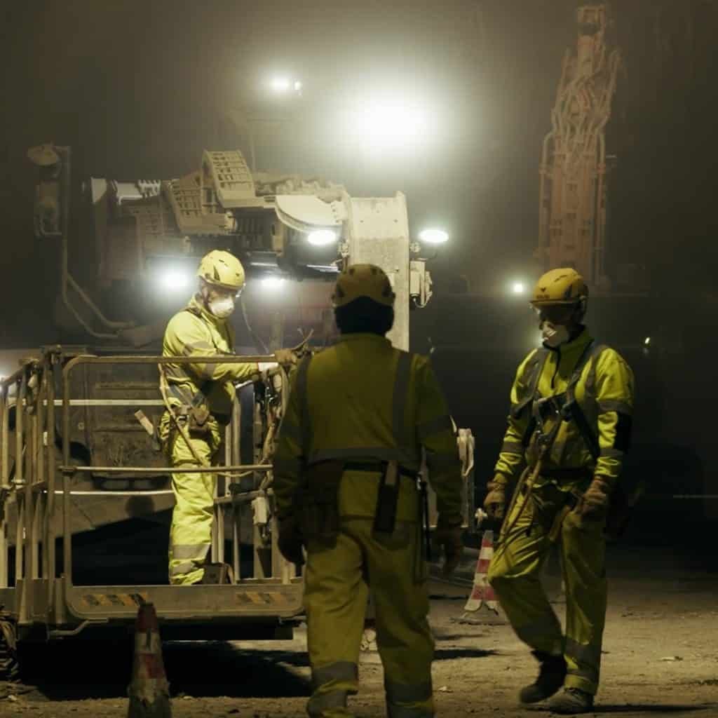 Three construction workers in safety gear and helmets efficiently tackle their tasks on a foggy night, silhouetted by bright lights and large machinery, as they diligently maintain the vital Autostrade highways of Italy.