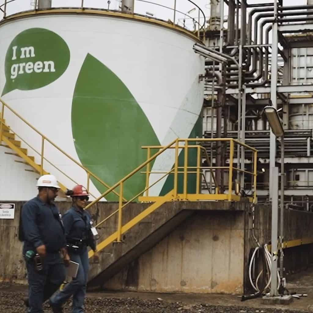 Two people in protective gear walk past an industrial tank with "I'm green" painted on it and large green leaf graphics.