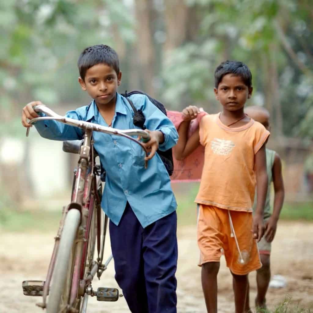 Three children stroll along a dirt path; the boy in the foreground, in a blue shirt, pushes a bicycle. Alongside him is another boy in orange, chatting excitedly about the new center dedicated to affordable water solutions. A third child follows closely behind.