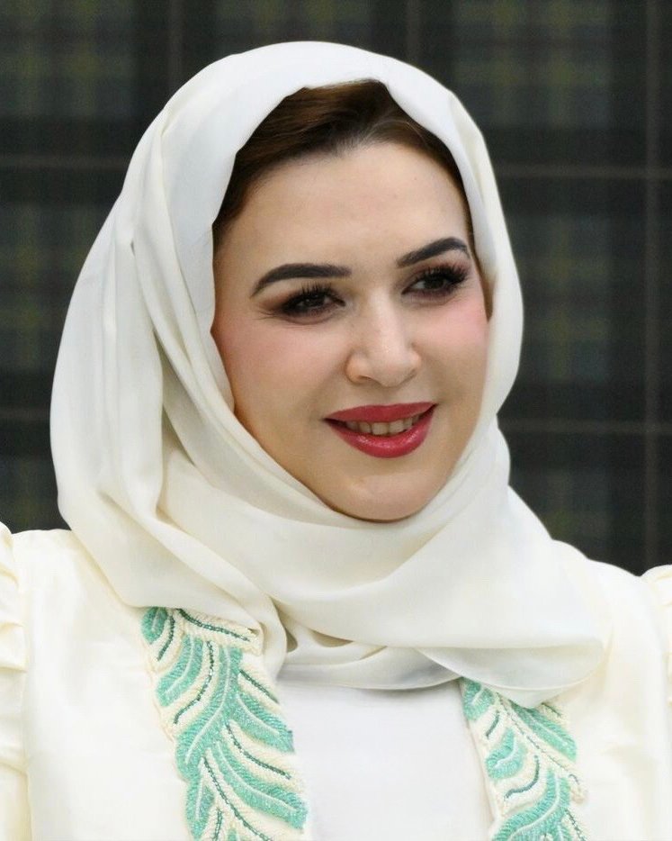 Dr. Nahla Al Qassimi, wearing a white headscarf and an embroidered top, smiles warmly against a dark, patterned background.