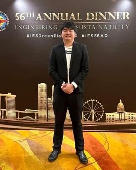 Deckson Chun Sin Ang, in a black suit, stands confidently before a backdrop adorned with "56th Annual Dinner, Engineering Sustainability" text and cityscape illustrations.