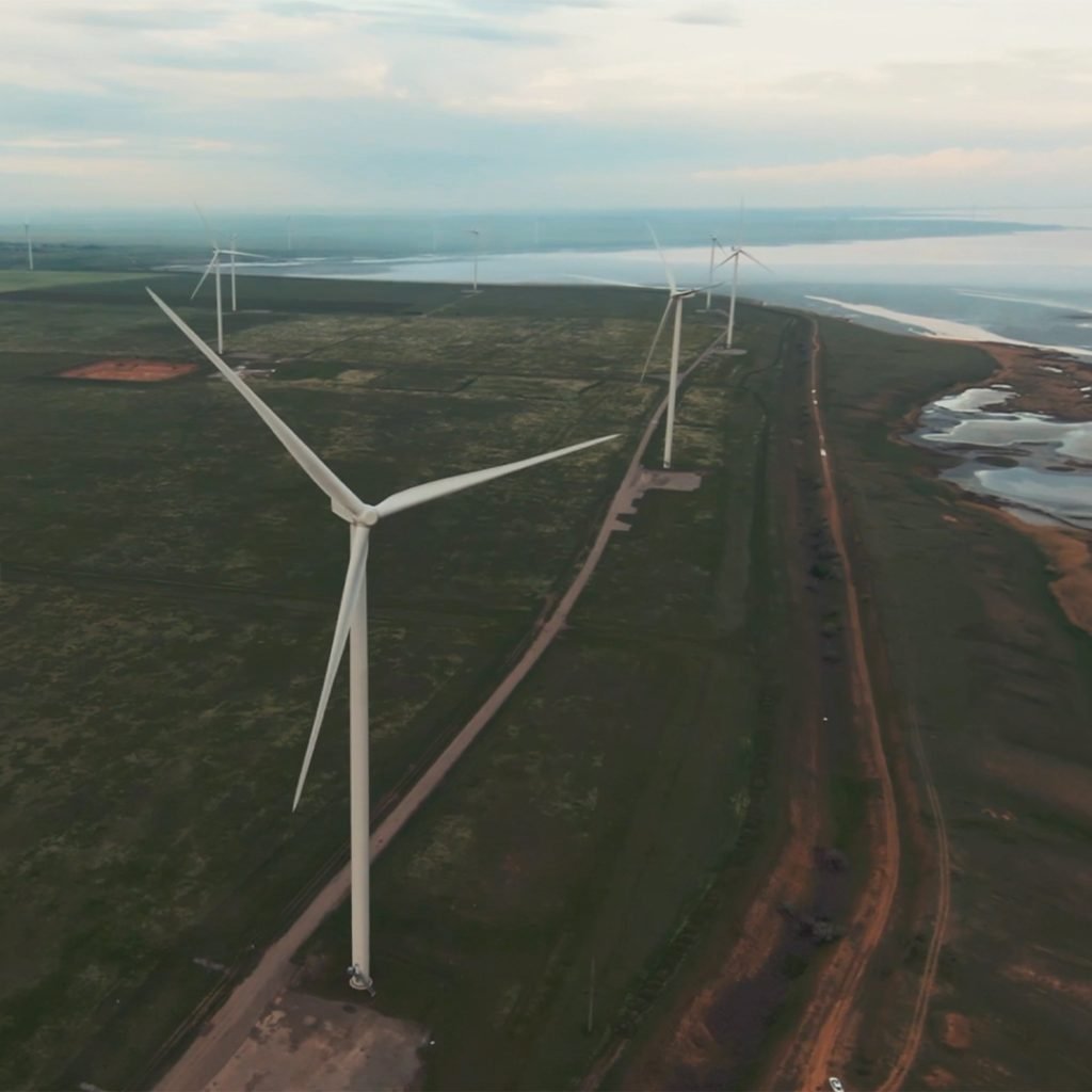 Wind turbines on a grassy landscape with a dirt road and water in the background, under a cloudy sky.
