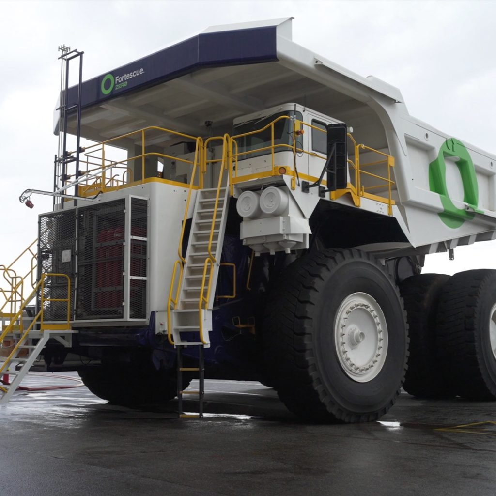 Large mining truck with Fortescue logo, parked on a wet surface. The truck has a white body, large tires, and yellow railings. Steps lead to the driver's cab. Overcast sky in background.