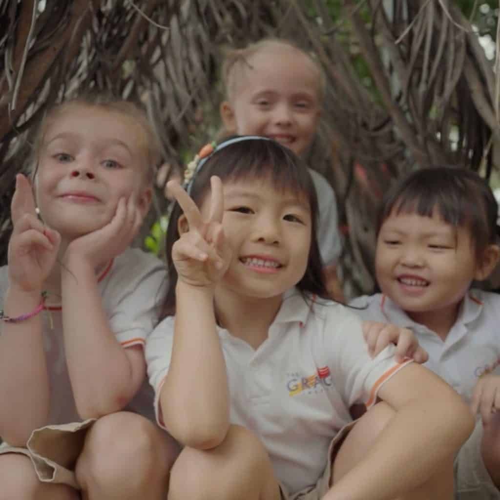 Four children from The Grange Institution smile and make peace signs as they sit together under a lush canopy of leaves.