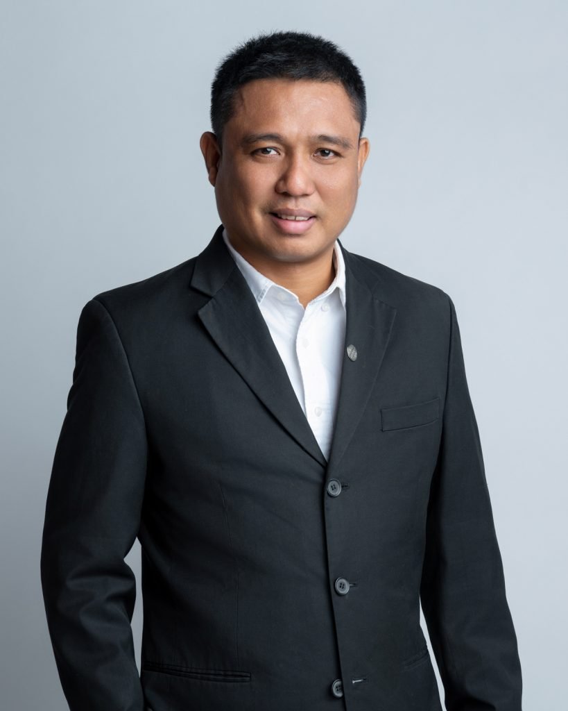 Habibie Razak, in a black suit and white shirt, stands poised against a plain background.