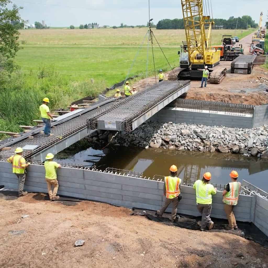 Construction crew in safety vests and helmets installing a bridge over water using cranes, with workers guiding beams into place on a sunny day.