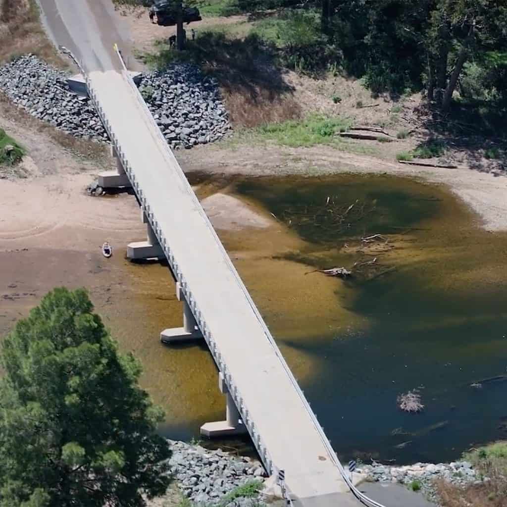 A narrow concrete bridge spans a shallow, partially dried river with exposed rocks and debris. A person stands near the water's edge. Trees and a vehicle are visible in the background.