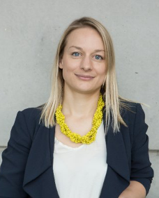 Katie Cresswell-Maynard, with shoulder-length blonde hair, dons a black jacket and white top. Her bright yellow necklace adds a vibrant touch as she stands against a subdued gray background.