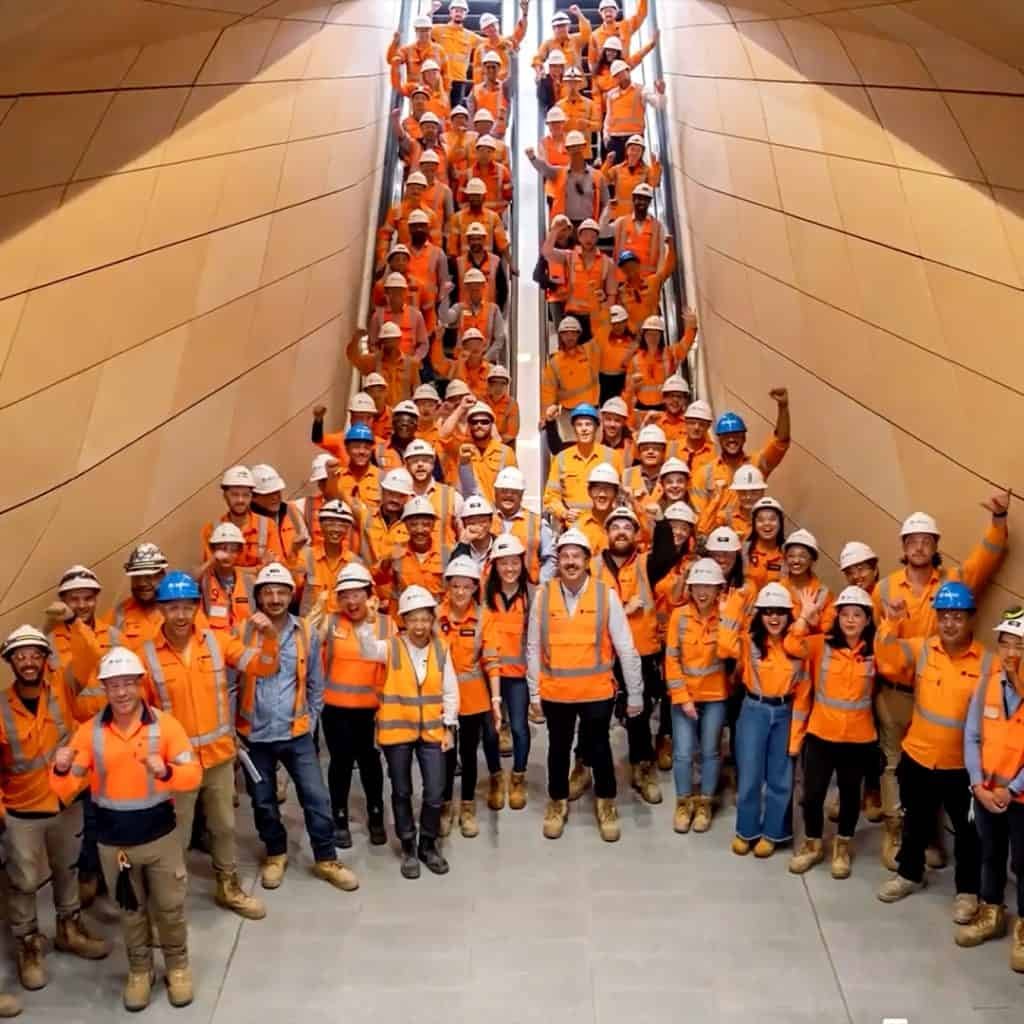 A team of construction workers from Laing O’Rourke, clad in orange vests and helmets, pose on the stairs, creating a perfectly organized assembly.