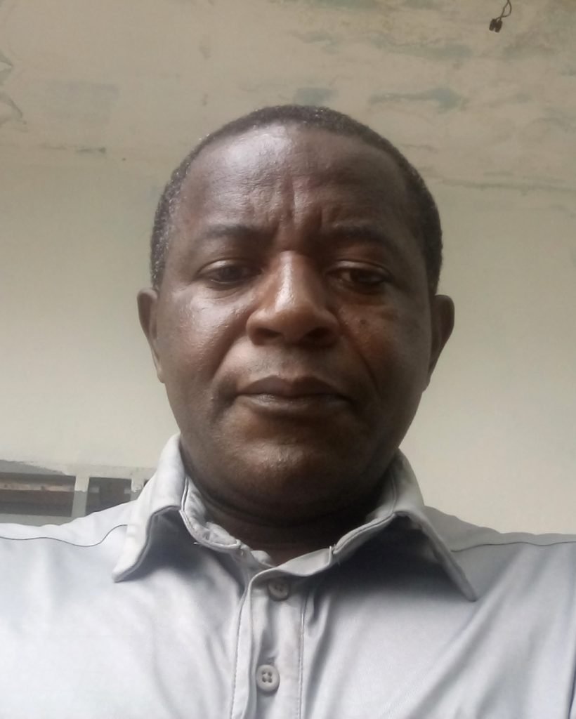 A person wearing a button-up shirt looks directly at the camera with a neutral expression, reminiscent of Norbert Tchiadje Tchouaffe. The background is plain and partially visible.