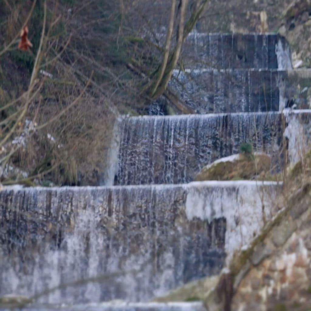A series of stepped waterfalls cascading down a rocky terrain surrounded by bare winter trees.