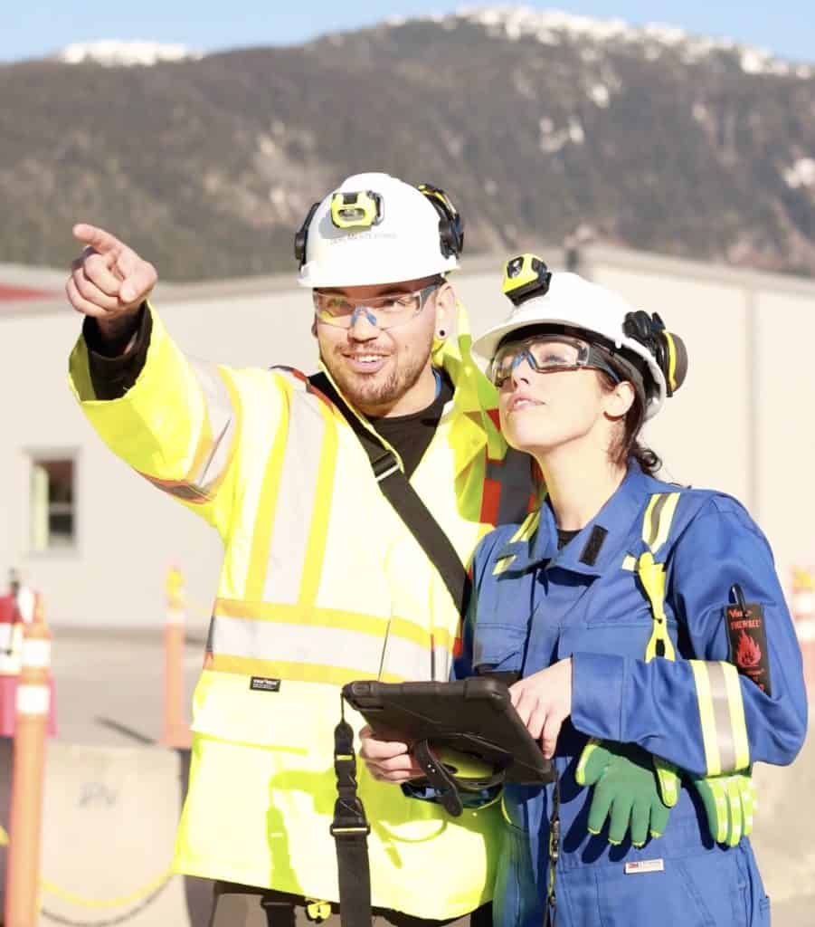 Two workers in safety gear and helmets are outdoors. One is pointing while the other holds a tablet, utilizing Kongsberg Digital technology. Snow-capped mountains form a stunning backdrop.