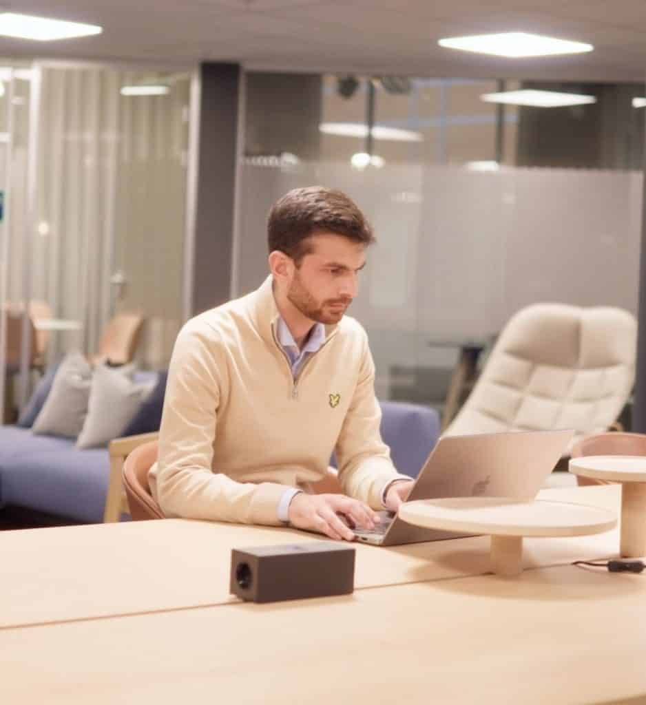 A man in a beige sweater is sitting at a desk, immersed in his laptop, surrounded by the sleek ambiance of a modern Kongsberg Digital office space.