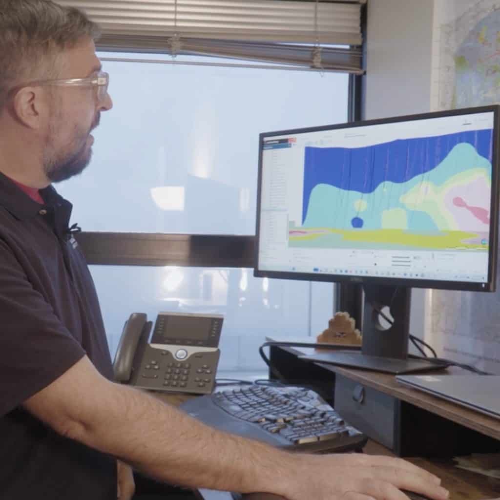 A man in glasses observes a computer screen displaying a colorful, abstract chart. An office phone and other equipment are on the desk beside him.