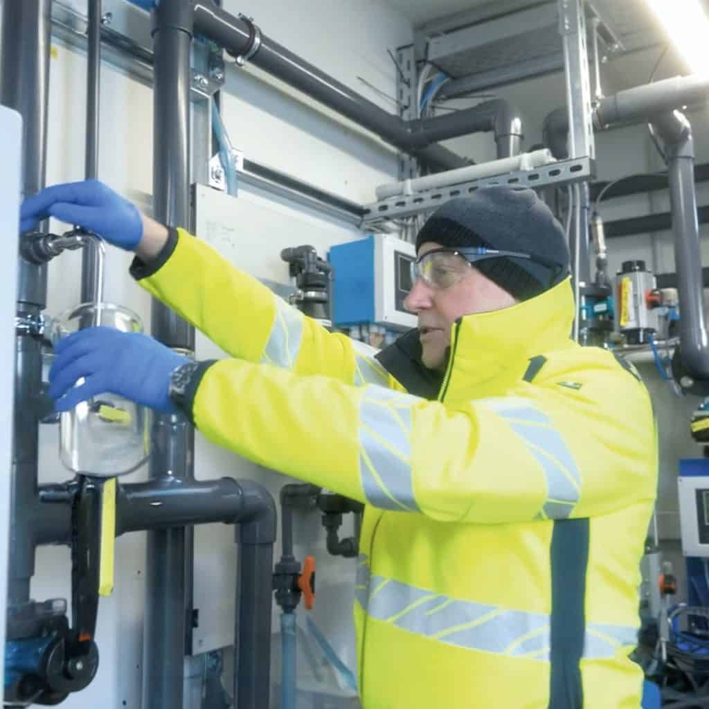 A person in a fluorescent jacket and gloves adjusts pipes in an industrial setting.