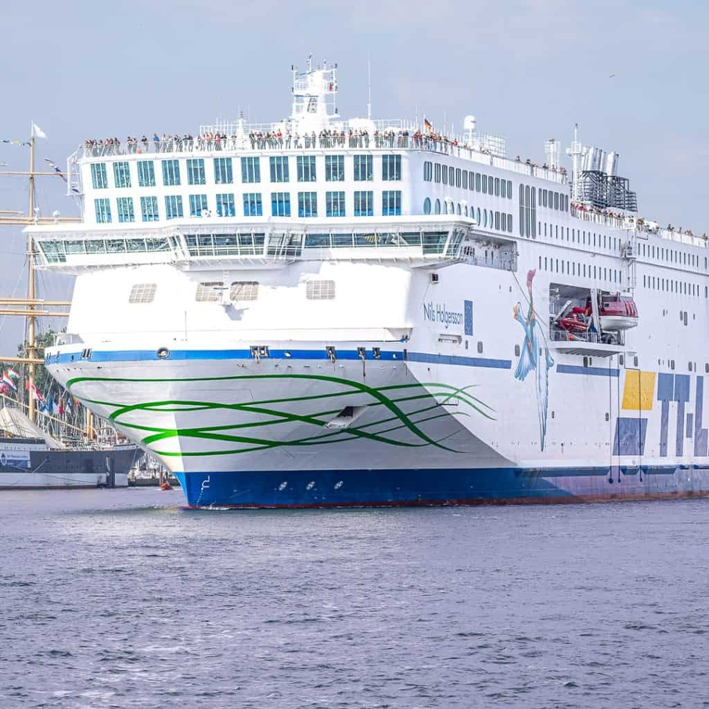 Large white ferry with green and blue stripes docked in a harbor, passengers on the upper deck, with a smaller ship visible in the background.